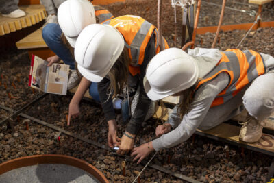 3 personnes casquées d'un casque de chantier, l'une tenant une fiche explicative et pointant un élément au sol, les deux autres accroupies, regardent dans la direction pointées - Agrandir l'image 2 sur 9, fenêtre modale